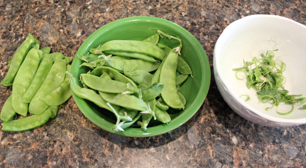 Snow peas in a green bowl wait to have the strings removed. Additional snow peas sit nearby with the strings already removed.