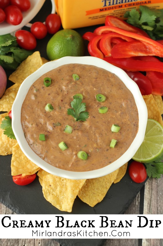 White bowl of black bean dip. Boil is surrounded by chips and veggies for dipping.