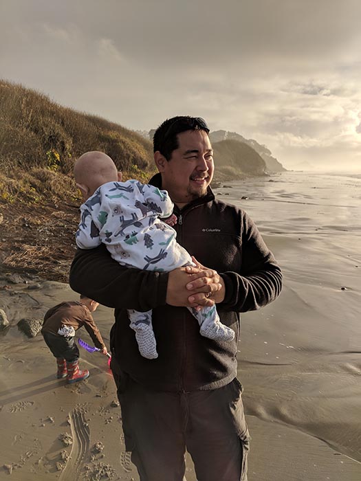 Baby Ella in the Dad's arms on the beach at the Oregon Coast.  Jack is digging in the sand in the background.