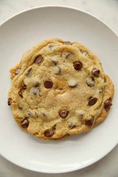 A bakery style large soft and chewy chocolate chip cookie sits by itself on a white plate. You can see some creamy peanut butter peaking through the cookie in the middle where it was stuffed with peanut butter.