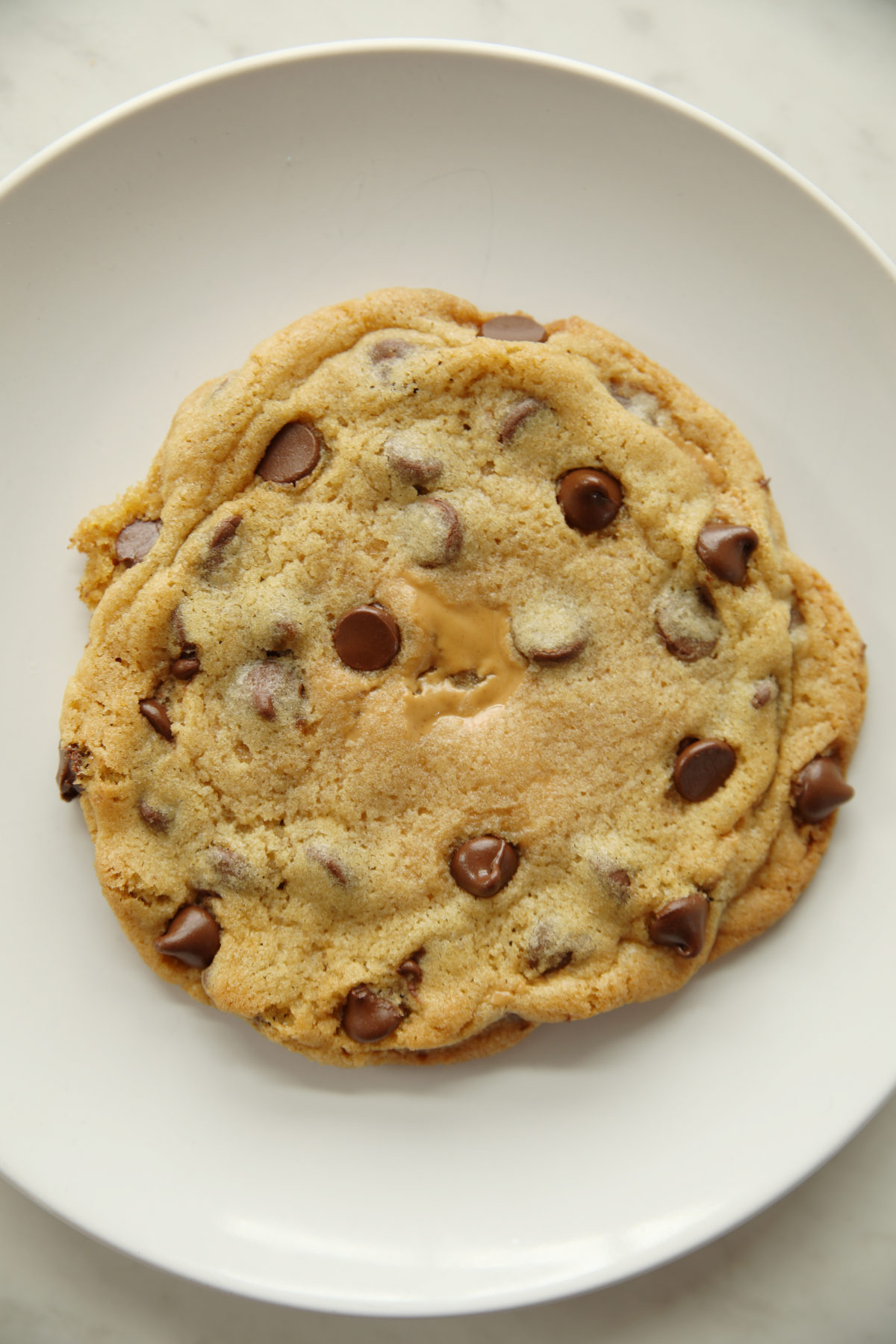 This is a photo of a large peanut butter stuffed chocolate chip cookie on a white plate.  You can see the peanut butter peaking through the middle of the chocolate chip cookie.  