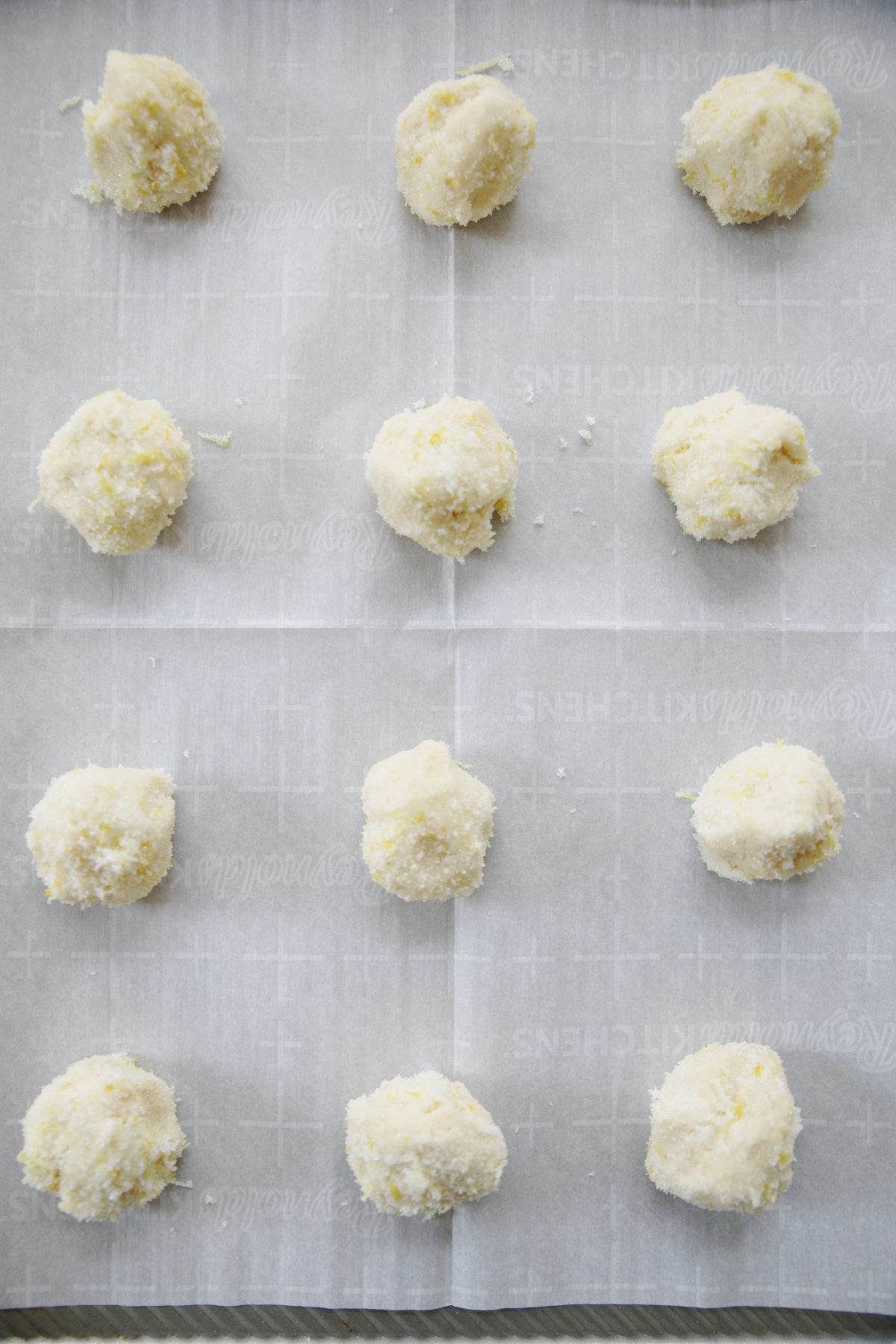 12 lemon sugar cookie dough balls are on a tray waiting to go into the oven. You can see the lemon sugar the balls are rolled on and a parchment paper liner for the baking tray.