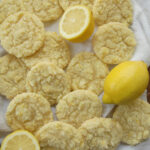 A big spread of soft, chewy lemon sugar cookies coated in lemon sugar are laid out on a white cloth. A few fresh lemons are also on the cloth.