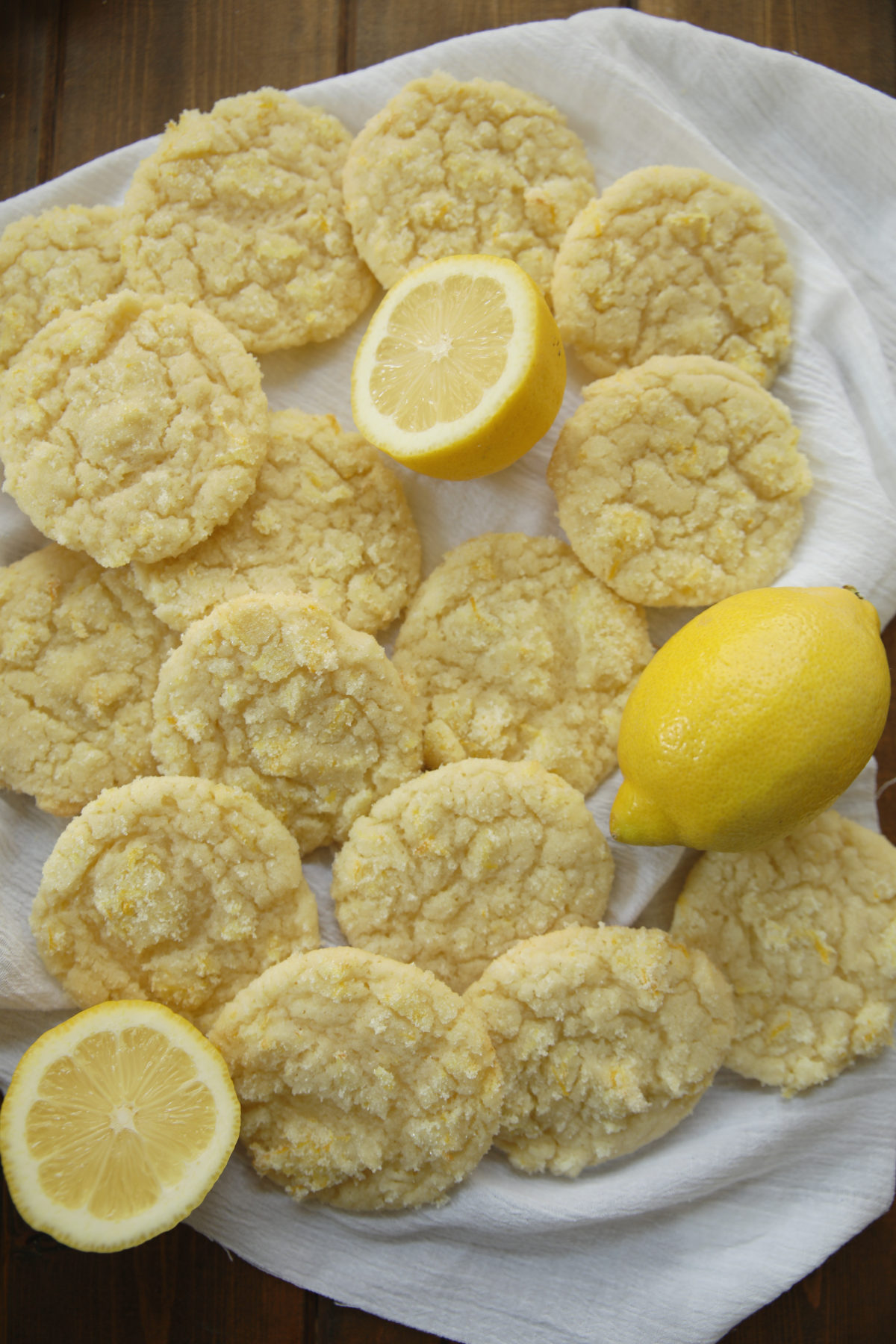 A big spread of soft, chewy lemon sugar cookies coated in lemon sugar are laid out on a white cloth. A few fresh lemons are also on the cloth. 