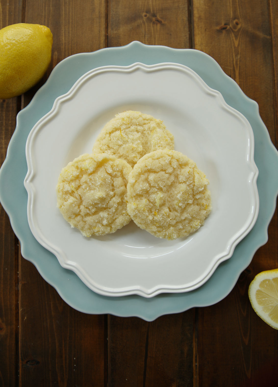 3 Lemon sugar cookies on a white plate stacked on a blue plate. You can see the lemon sugar on the cookies and some fresh lemons on the table by the plate